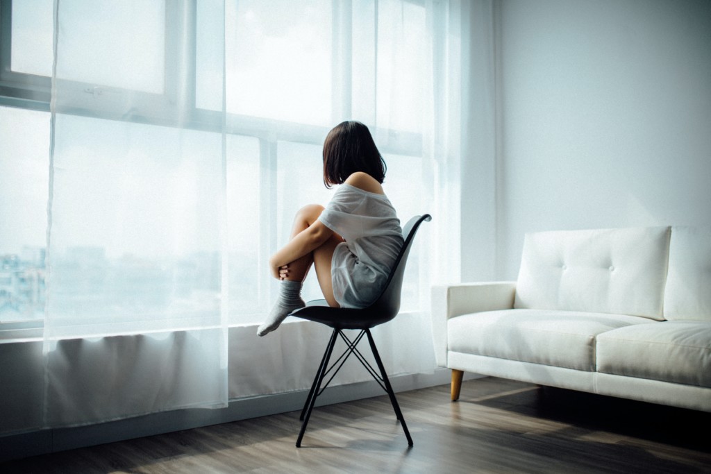 person with short dark hair sits on chair in room looking out of window