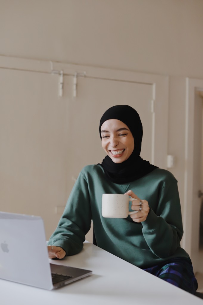 emotions displayed: happiness, contentment. woman sitting at a laptop, smiling with a white mug