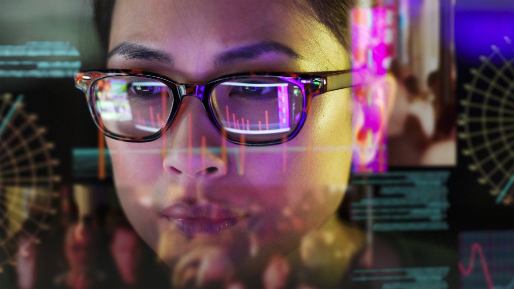 Close up of a woman carefully studying moving data on her computer screen, the screen is unusual as it is transparent and the camera is looking through the back of the screen.