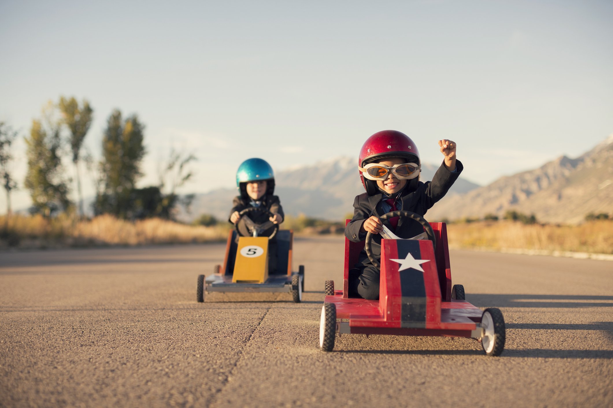 A young boy dressed as a businessman raises his arm in success as his homemade box car is in first place. Both boys are wearing helmets and goggles.