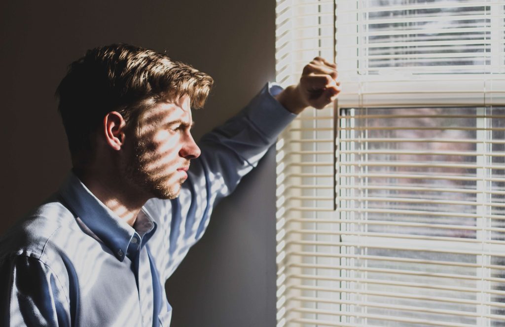 Stock image of a man looking out a window with blinds, casting shadow lines on him.